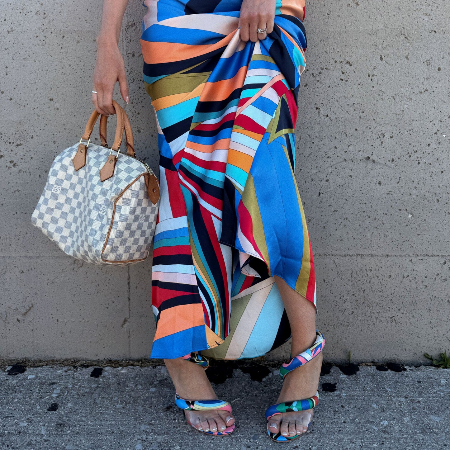 A person wearing a vibrant multicolor striped scarf and matching colorful flat sandals, holding a Louis Vuitton checkered handbag from North Shore Exchange against a concrete wall.