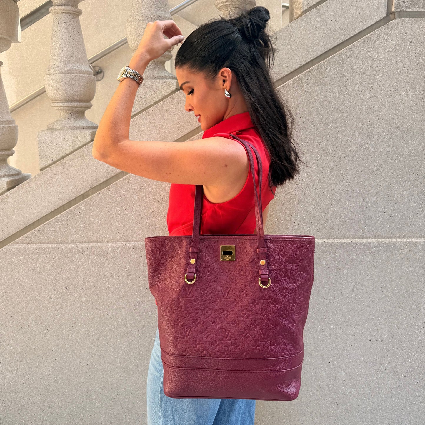 A woman in a red sleeveless top and light blue jeans stands on stone stairs holding a large burgundy monogram tote bag by Louis Vuitton from North Shore Exchange with gold hardware.