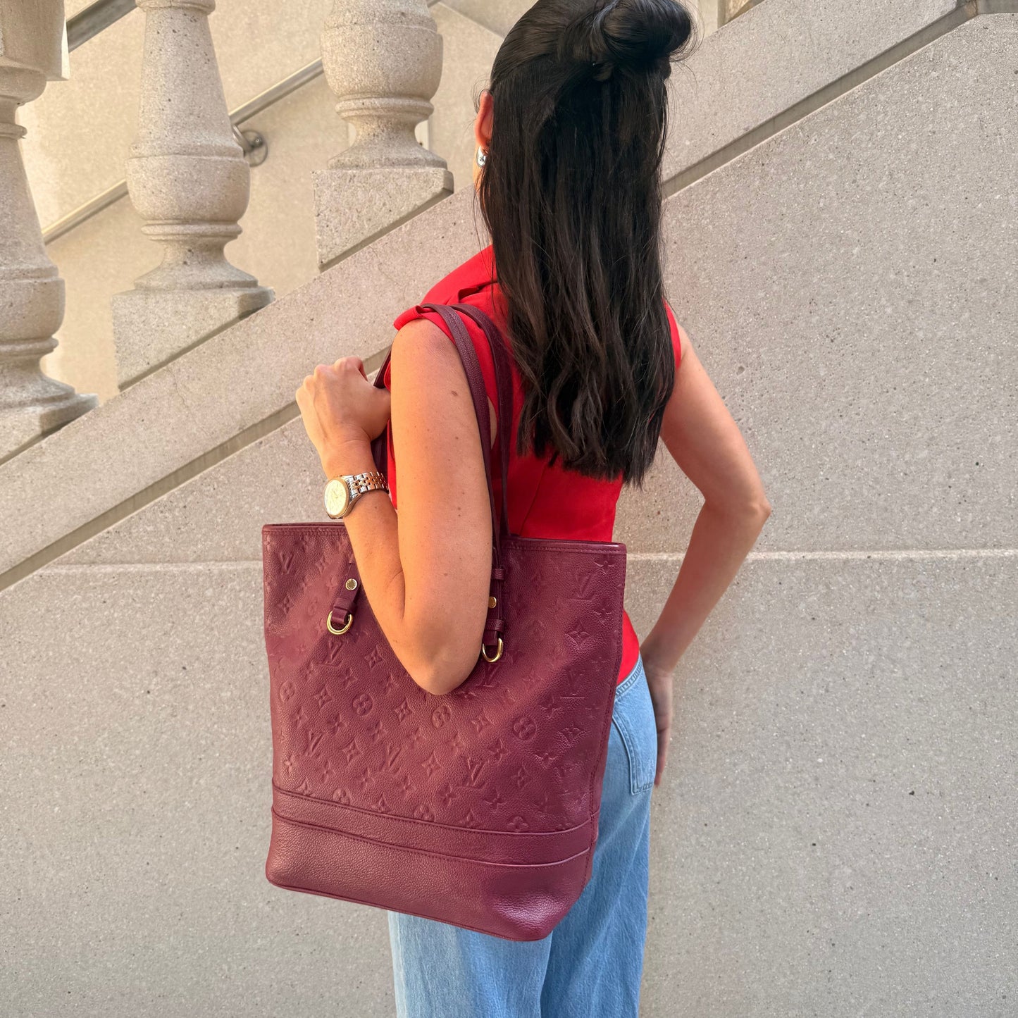 A woman viewed from behind wearing a red top and light blue jeans, carrying a large burgundy embossed leather tote bag by Louis Vuitton from North Shore Exchange on her shoulder against a light gray stone wall.