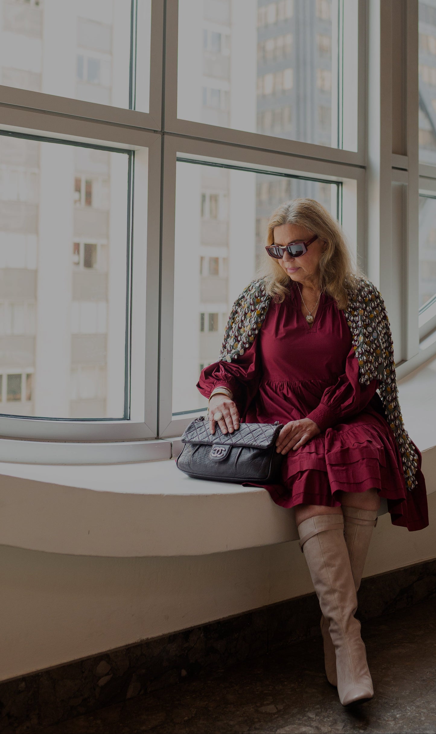 A woman sits on a window ledge wearing a burgundy long-sleeve ruffled dress, a patterned cardigan, beige knee-high boots, and holding a black quilted handbag.