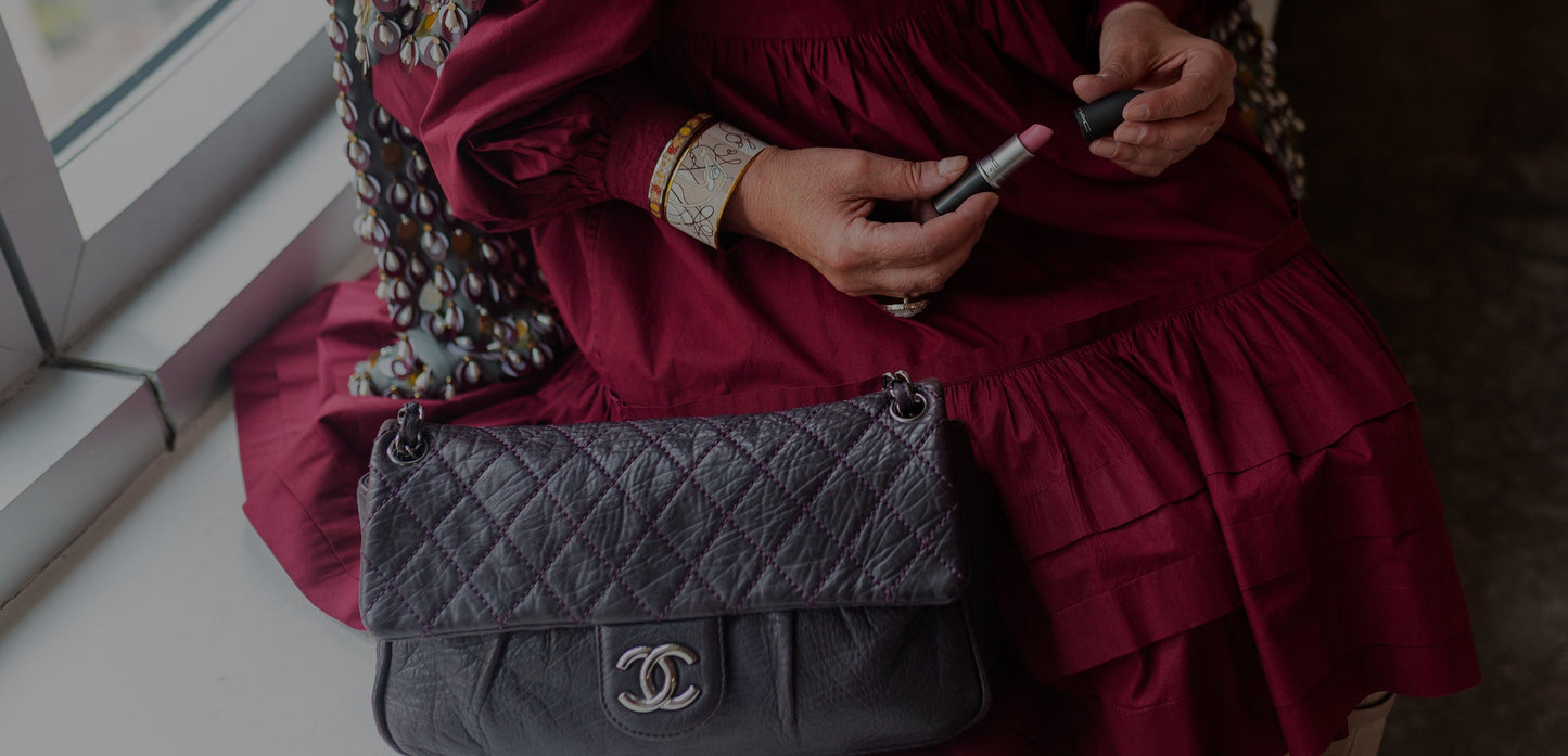 A person in a burgundy dress sits by a window, holding a pink lipstick while wearing a dark gray quilted shoulder bag with a silver interlocking CC logo, which indicates the brand Chanel.