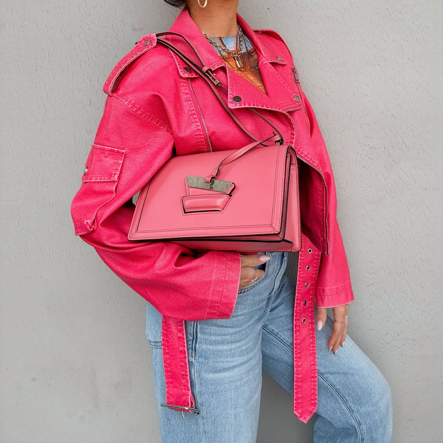 A person wearing a bright pink denim jacket and light blue jeans carries a structured pink handbag with black trim and a small metallic clasp by Loewe from North Shore Exchange against a light gray wall.