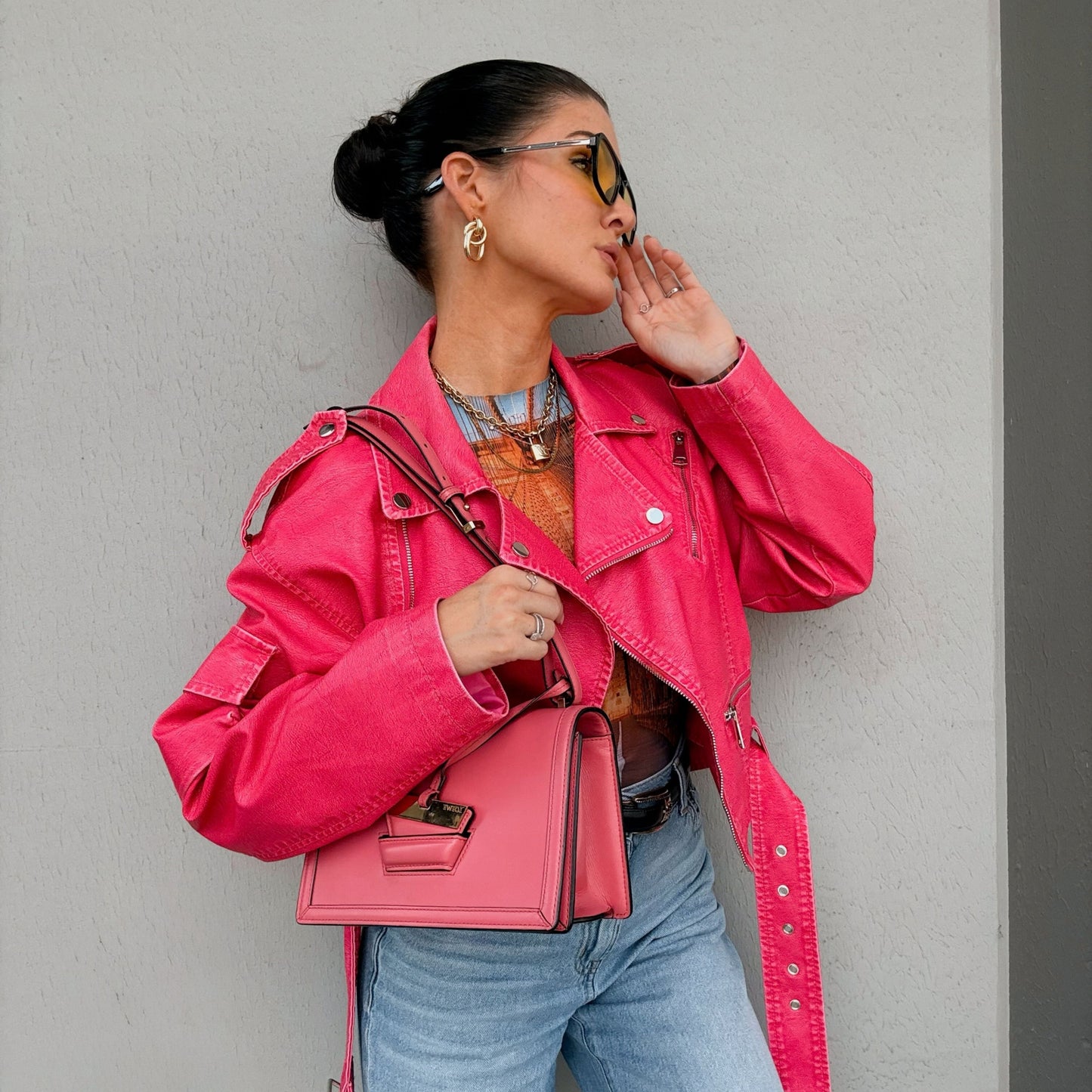A woman wearing a bright pink leather jacket and matching pink handbag by Loewe from North Shore Exchange, posing against a textured gray wall.